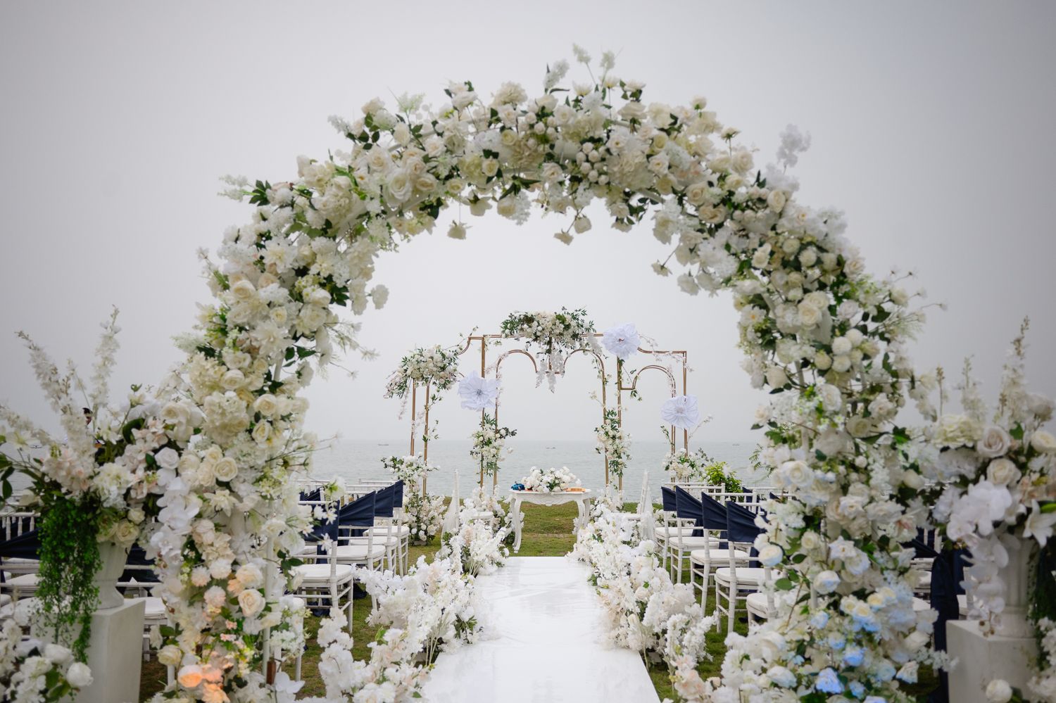 Elegant wedding ceremony aisle with white floral arches, lush blooms, and blue hydrangeas leading to a decorated altar.