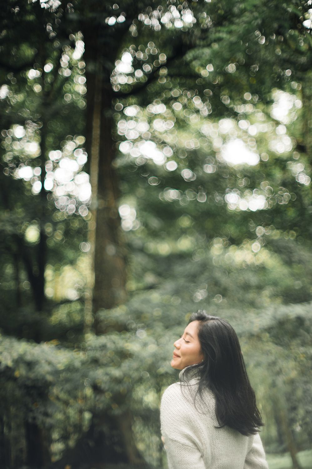Woman with eyes closed in peaceful meditation surrounded by lush green forest trees with bokeh light effect.