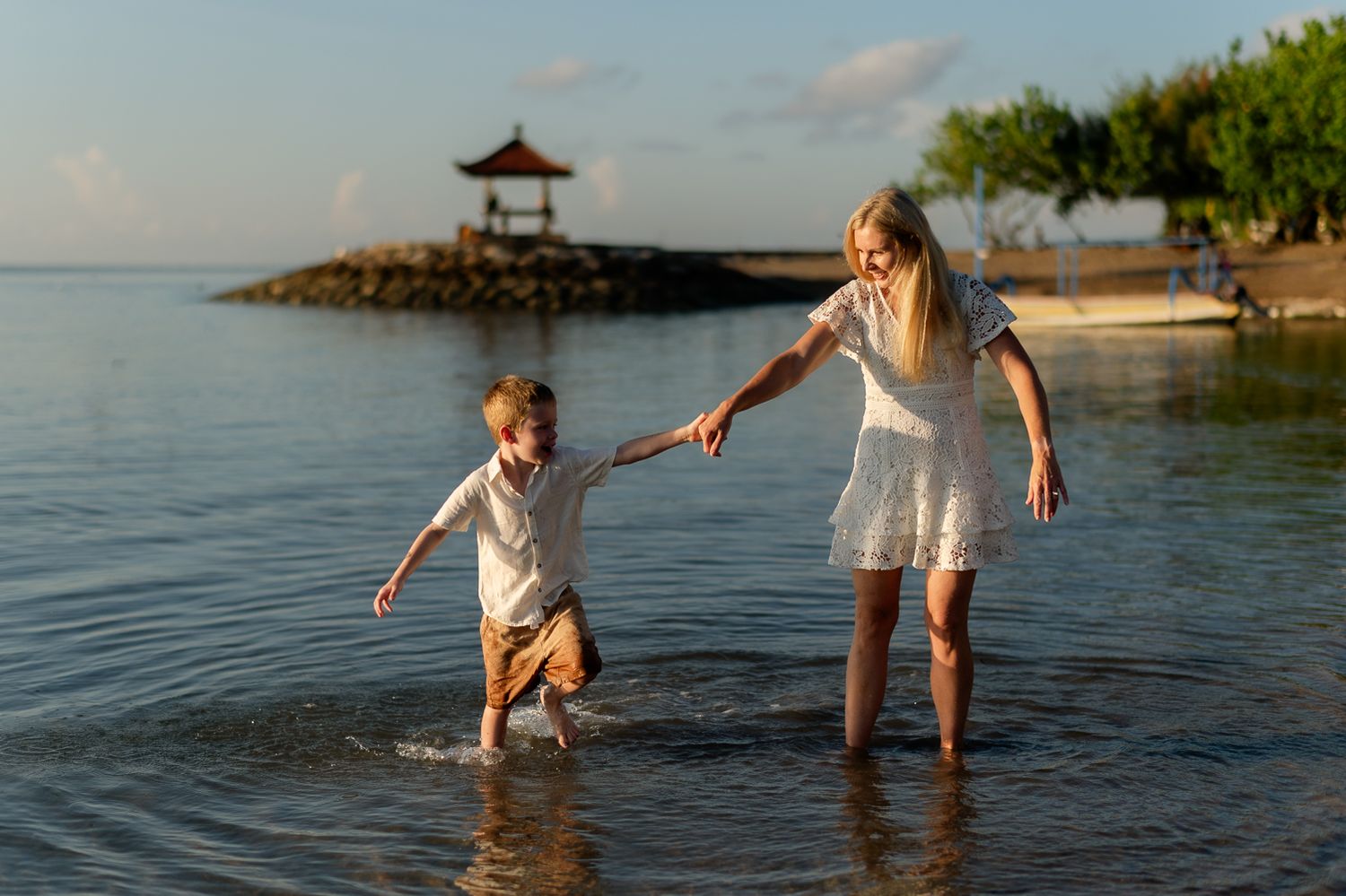 Mother and son laughing while holding hands and wading through shallow beach water at golden hour.