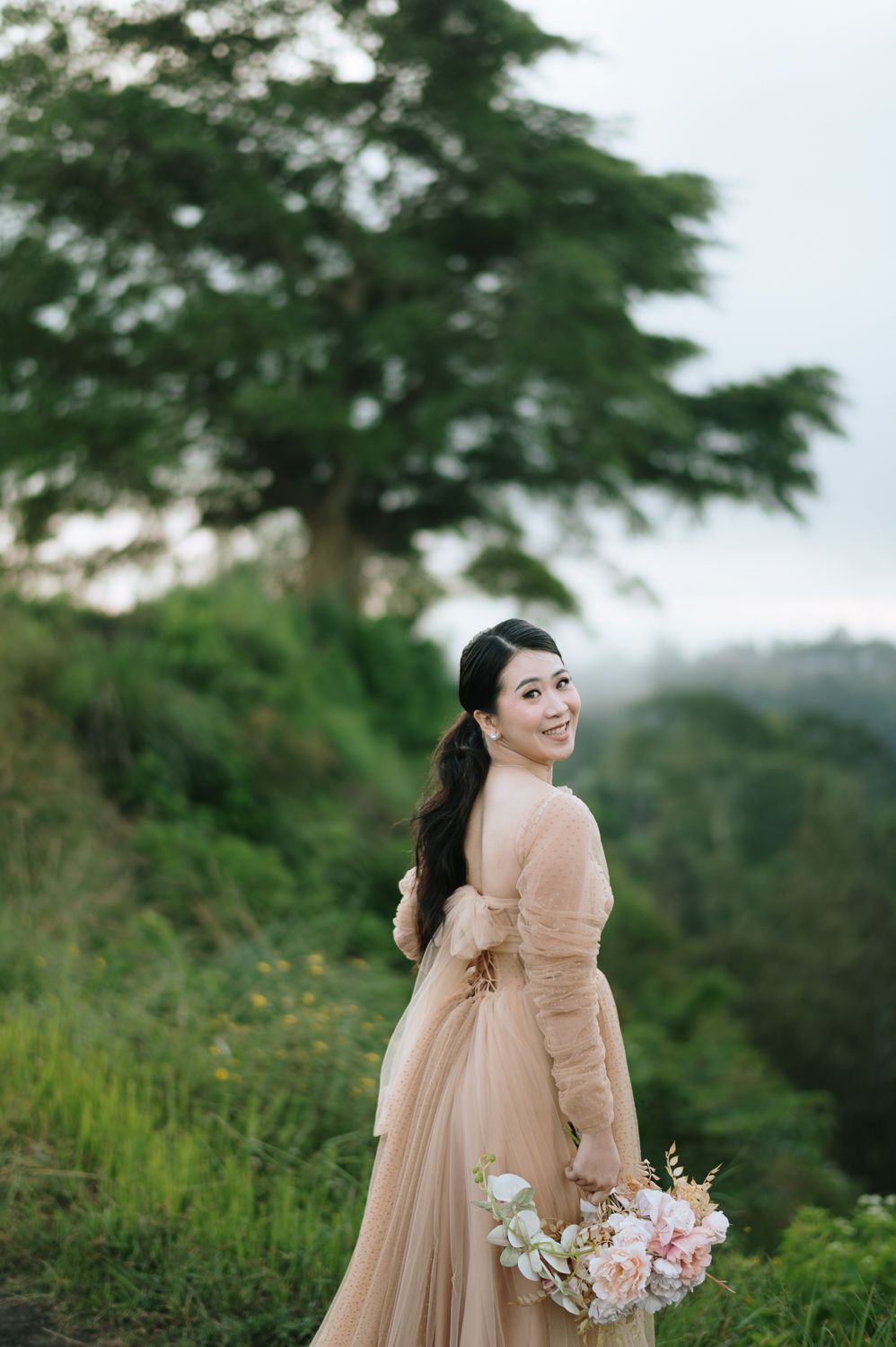 A smiling bride in a blush tulle gown holds a floral bouquet while posing outdoors among lush green hills.