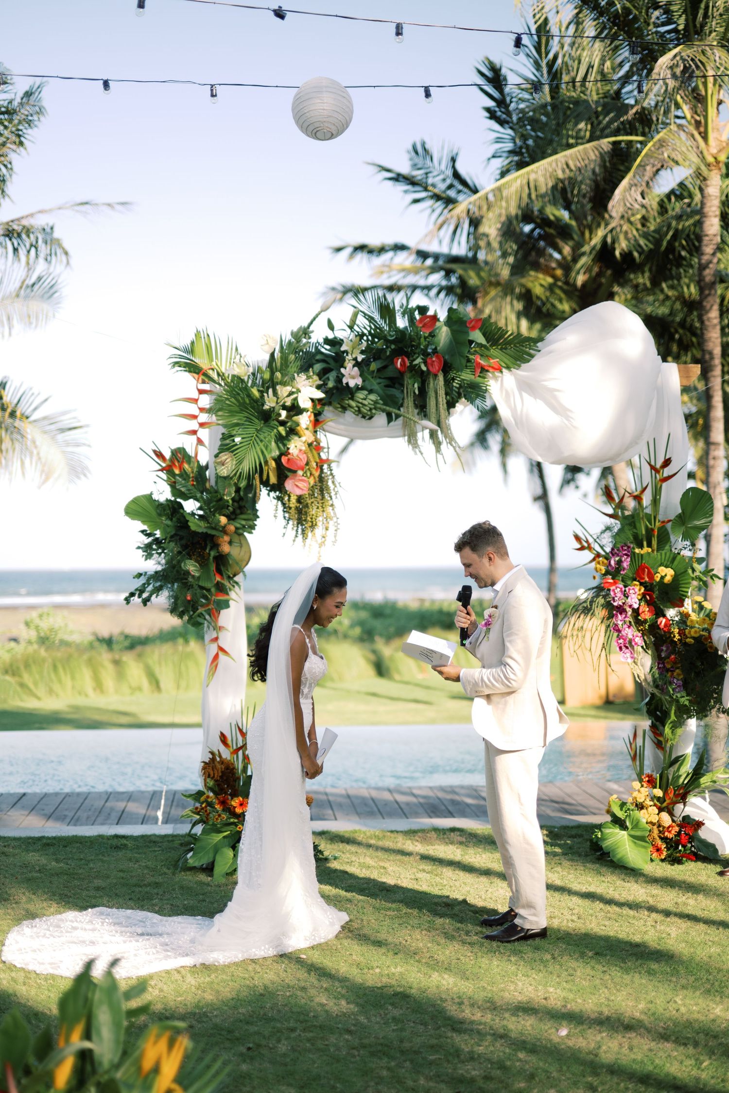 Bride and groom exchanging vows under a tropical floral arch with ocean backdrop and string lights at sunset.