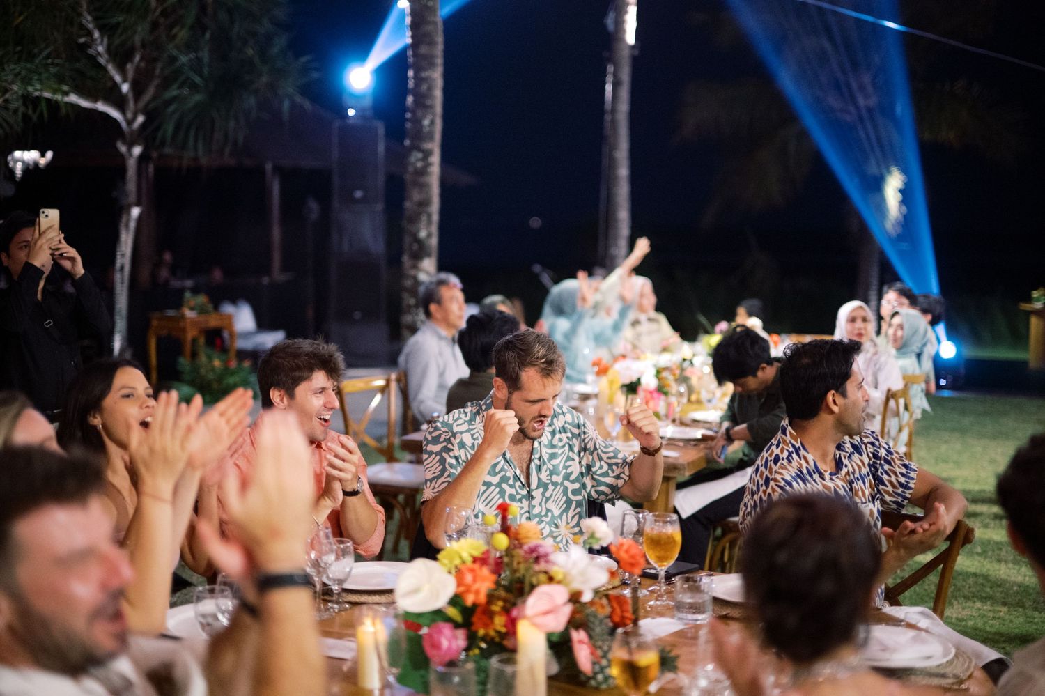 Guests clapping and celebrating at elegant outdoor wedding reception dinner under night sky lights.