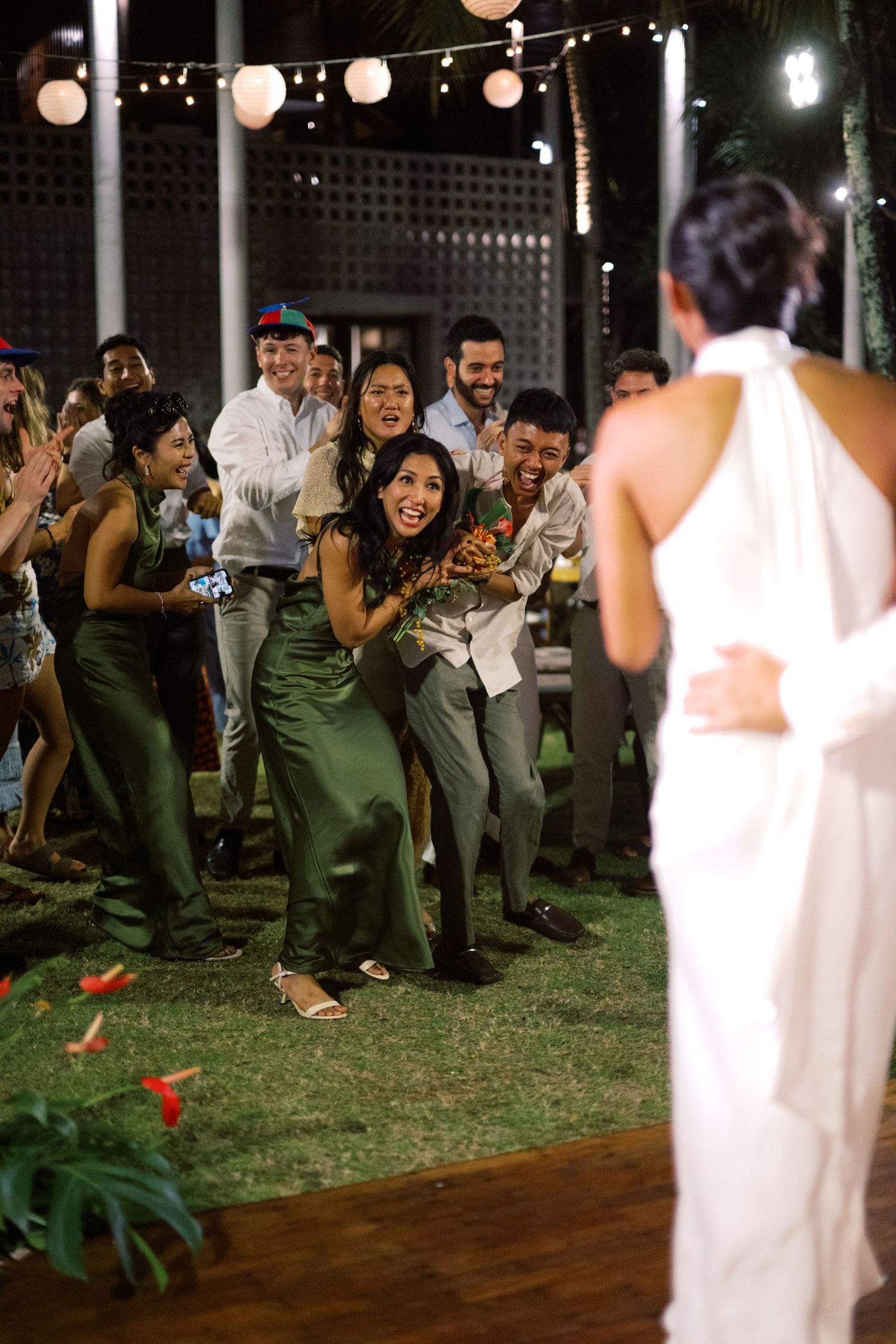 Excited wedding guests cheer as bride prepares to toss bouquet at tropical outdoor wedding reception.
