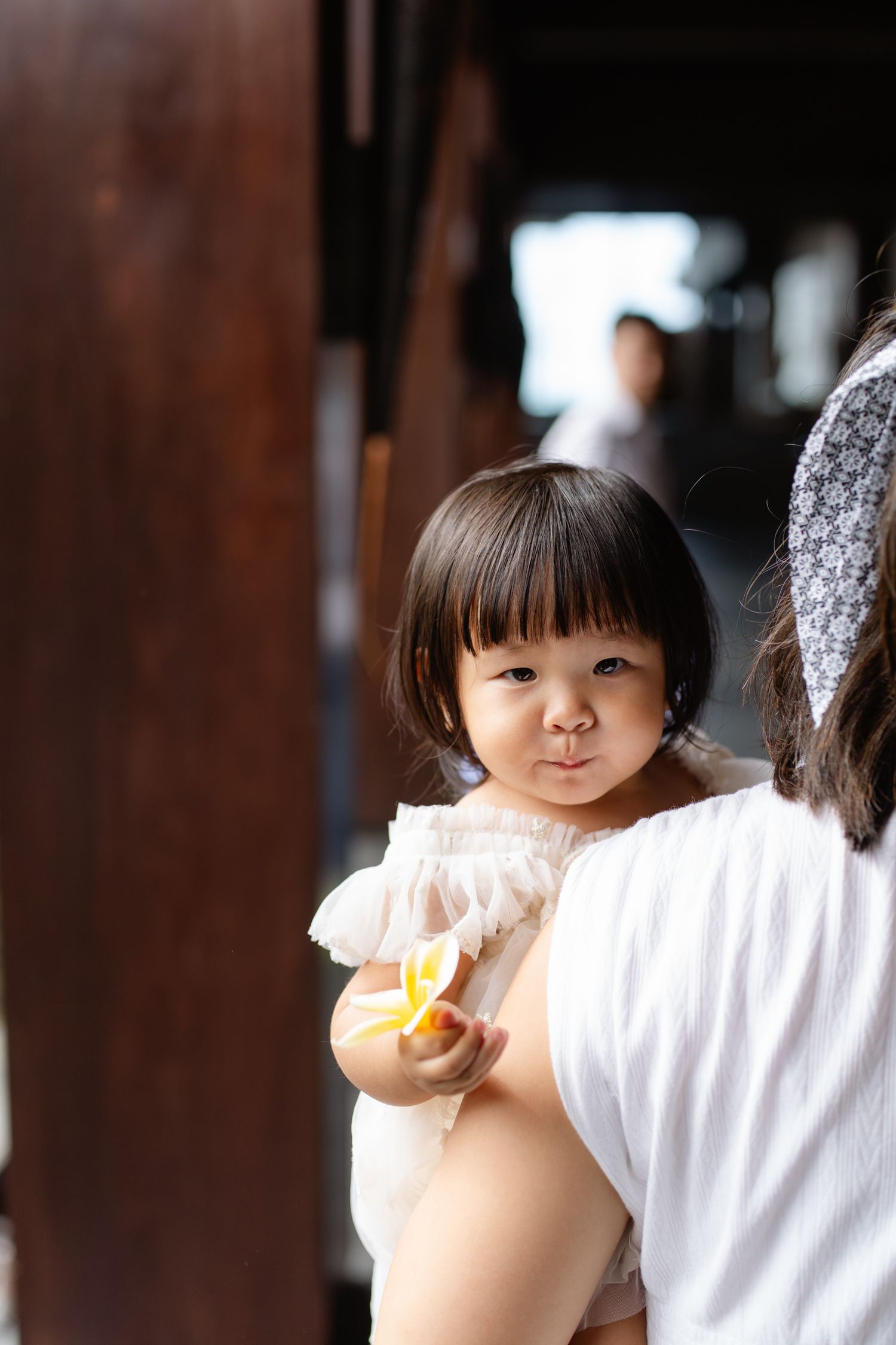 Adorable Asian toddler girl in white dress being held by mother, looking at camera while holding a yellow snack.