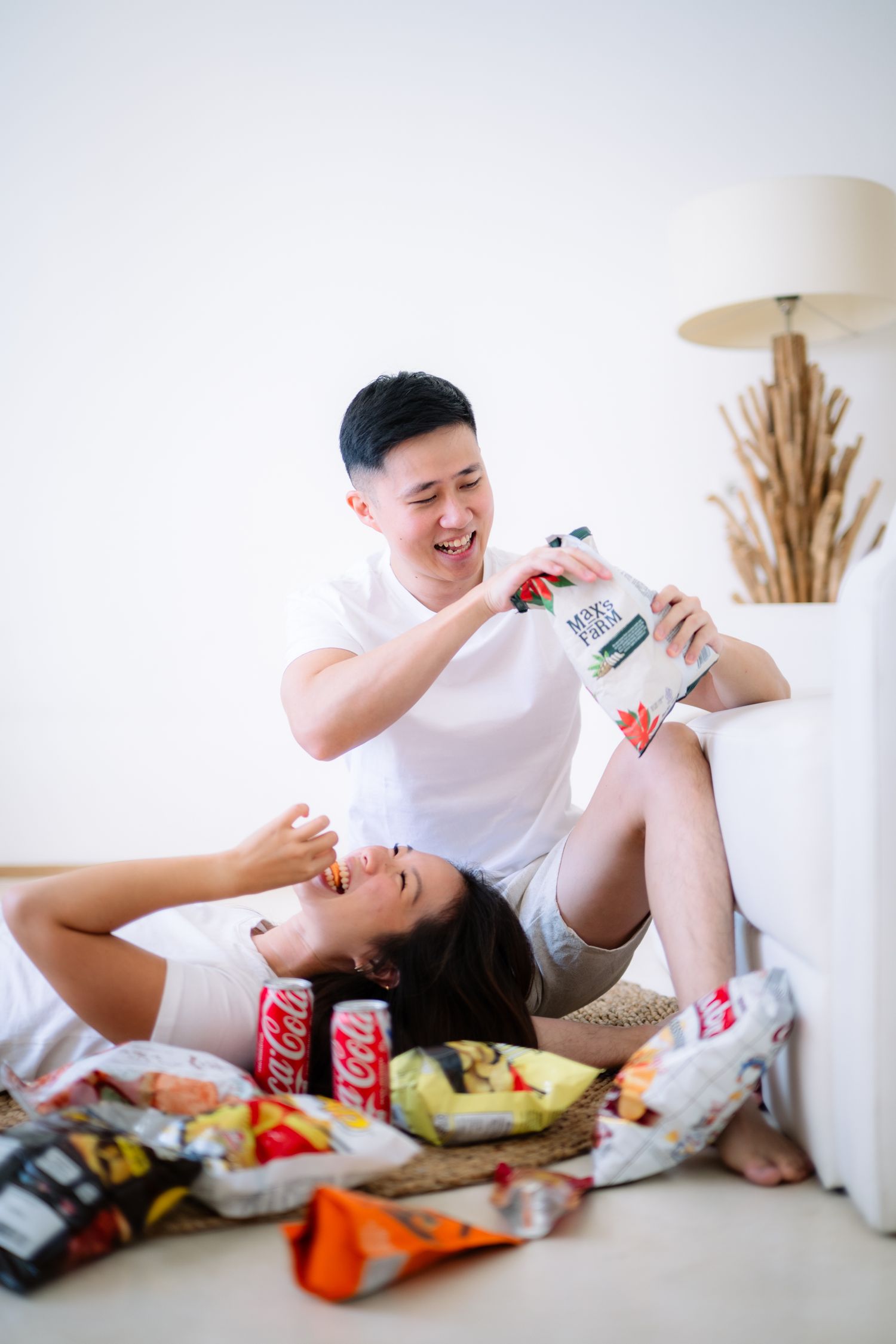 Young Asian couple laughing and snacking together on floor surrounded by chips and Coca-Cola cans.