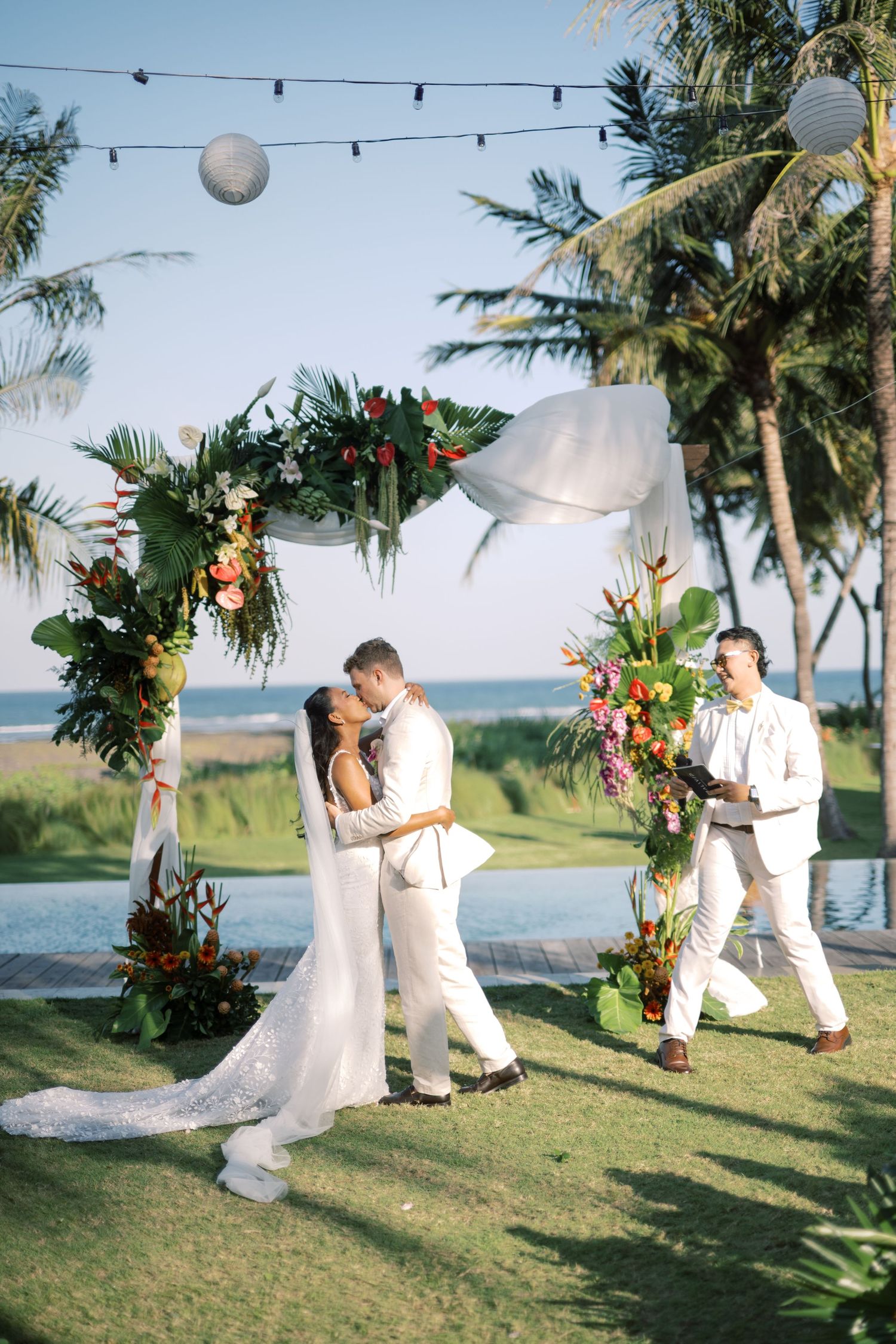 Couple shares first kiss at tropical outdoor wedding ceremony with floral arch, palm trees, and ocean backdrop.