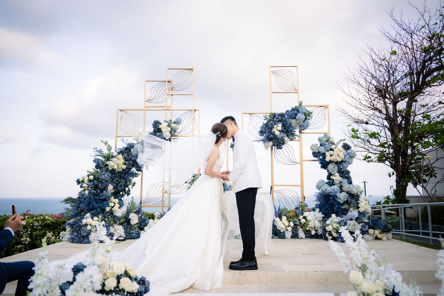 Bride in white gown and groom in black kiss during an outdoor wedding ceremony framed by blue and white floral arrangements and gold frames.