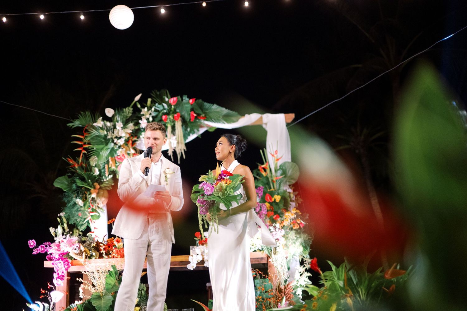 Couple in white attire stands at tropical floral altar during outdoor wedding ceremony at night.