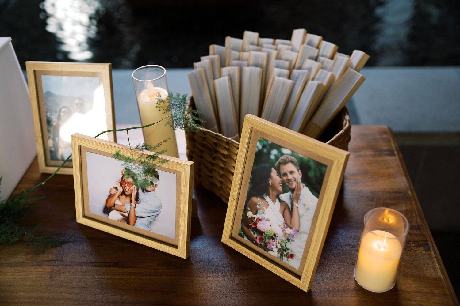 Gold-framed wedding photos displayed on wooden table with candles and basket of rolled programs at elegant reception.