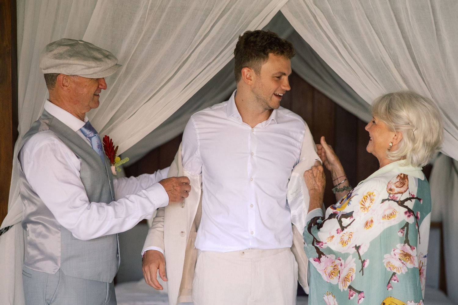 Elderly couple helping young groom dress in white shirt before wedding ceremony under draped canopy.