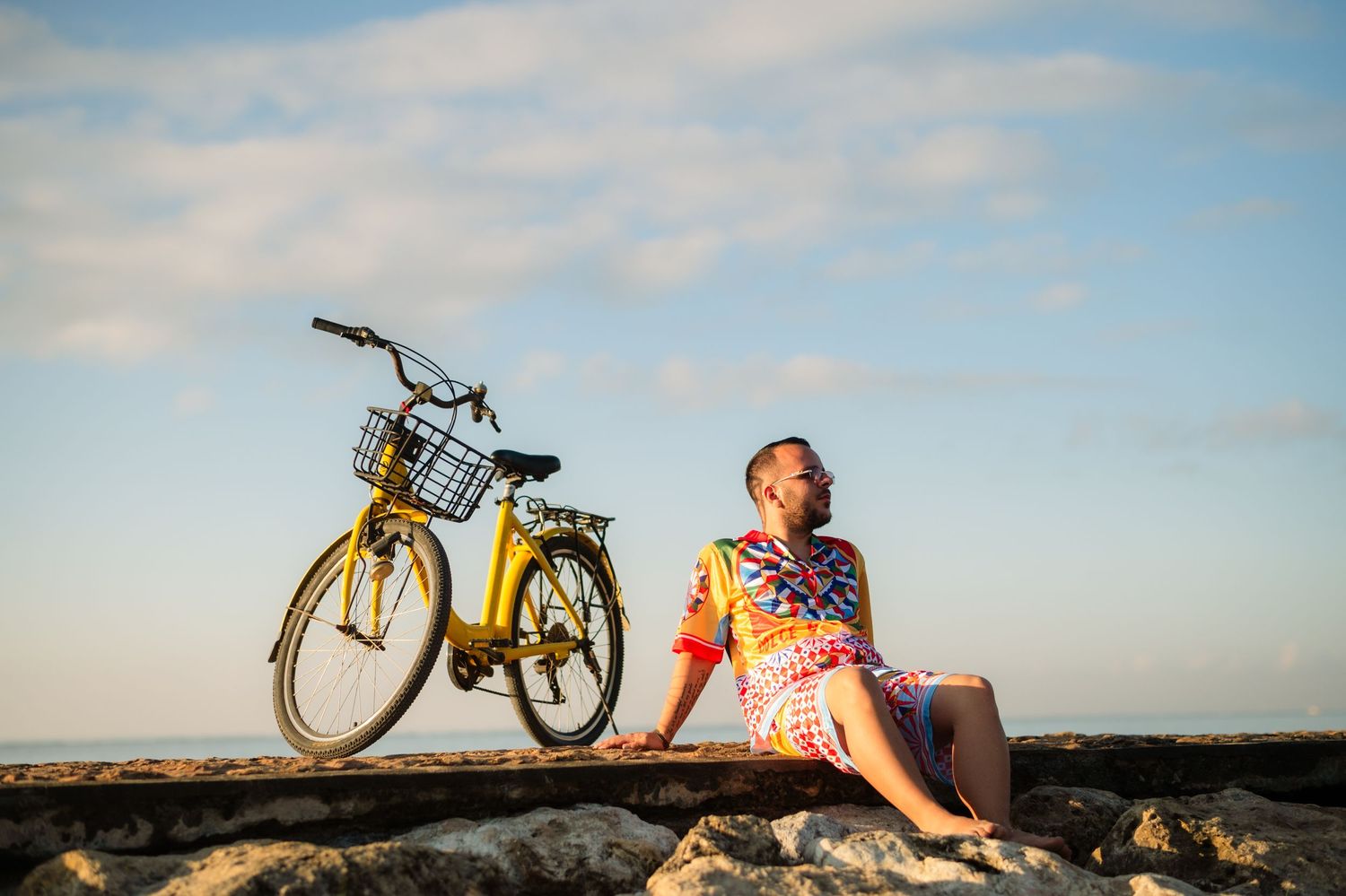 Woman in colorful dress sits on rocks beside a yellow bicycle with basket, enjoying a sunny coastal view.