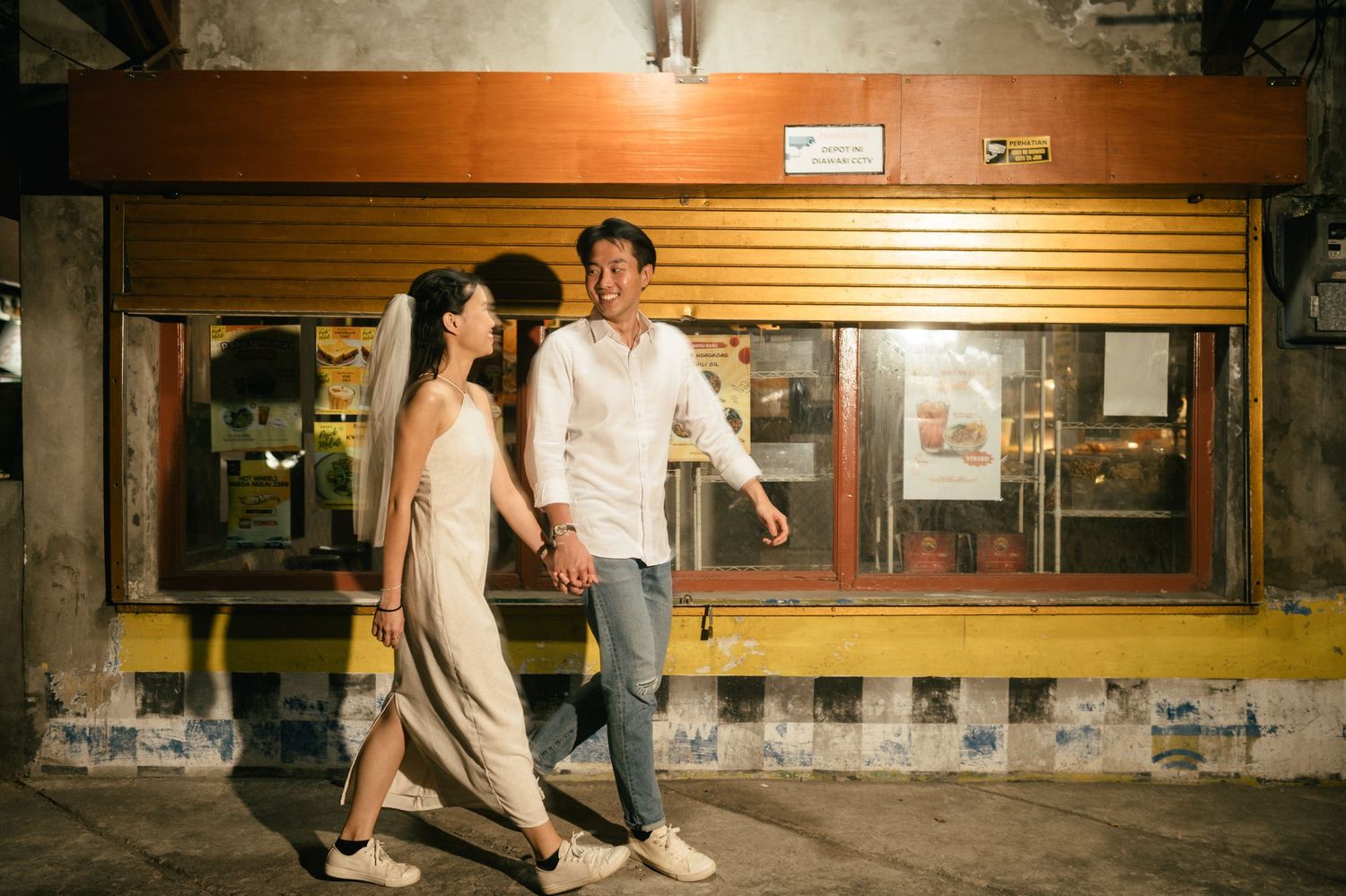 Couple walking hand in hand past a vintage storefront at night, smiling and dressed in casual elegant attire.