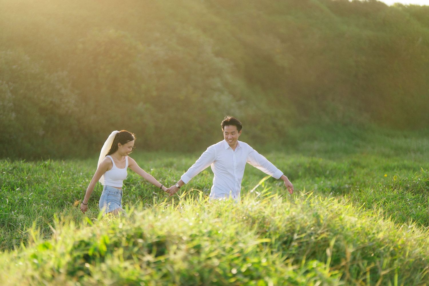 A happy couple holds hands while walking through a sunlit green meadow together.