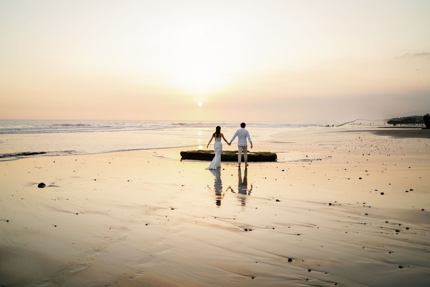 Bride and groom holding hands facing the ocean sunset on a serene reflective beach.