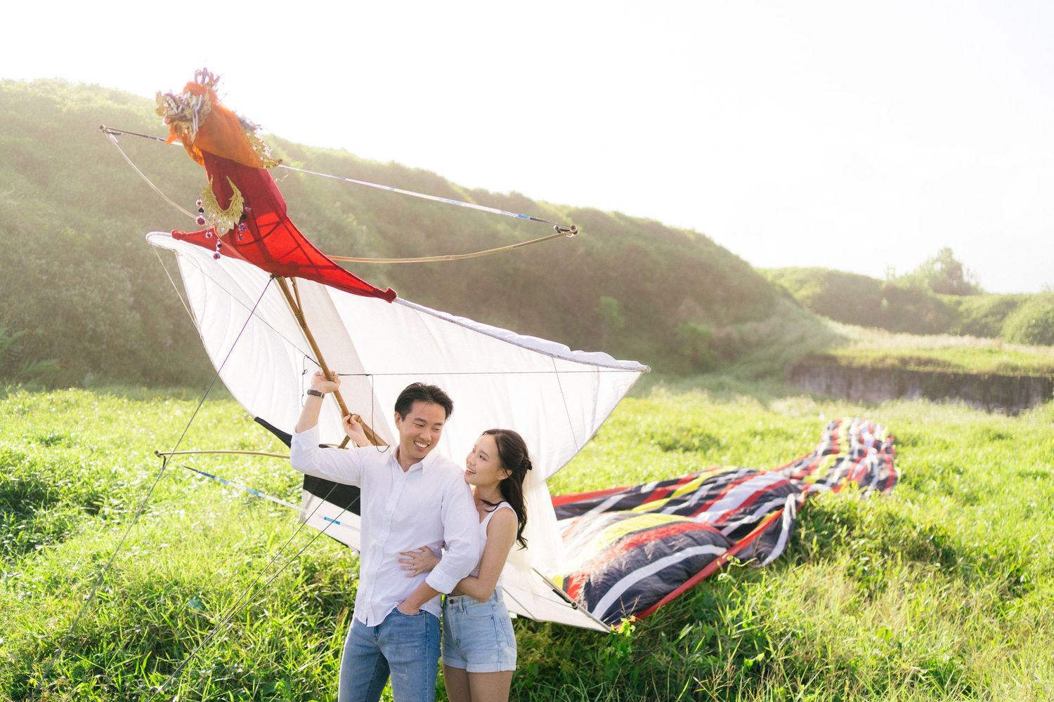 Couple smiling and flying a large colorful kite together in a sunny green meadow.