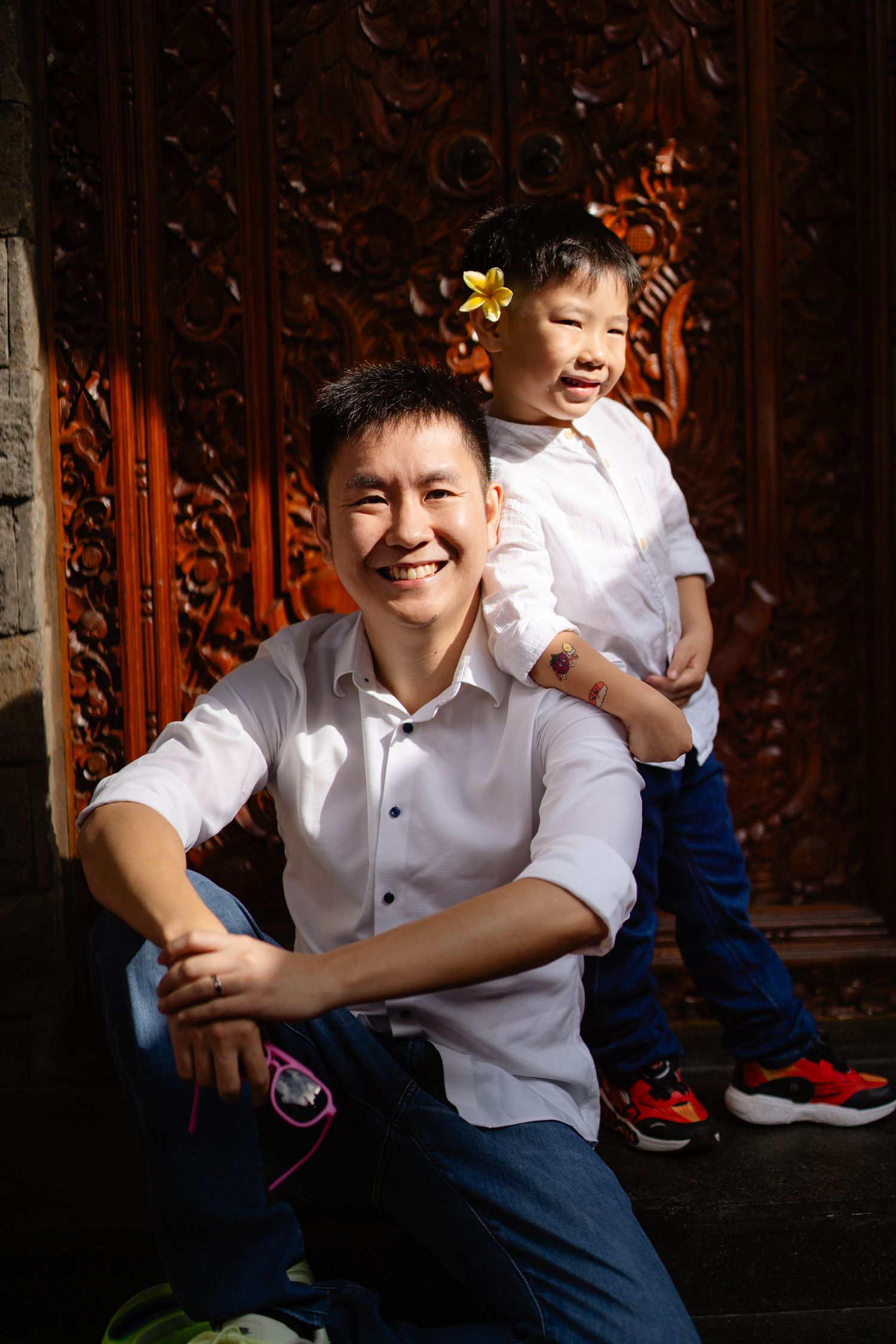 Smiling Asian man sitting with cheerful young child wearing flower hair clip posed in front of ornate carved wooden door.