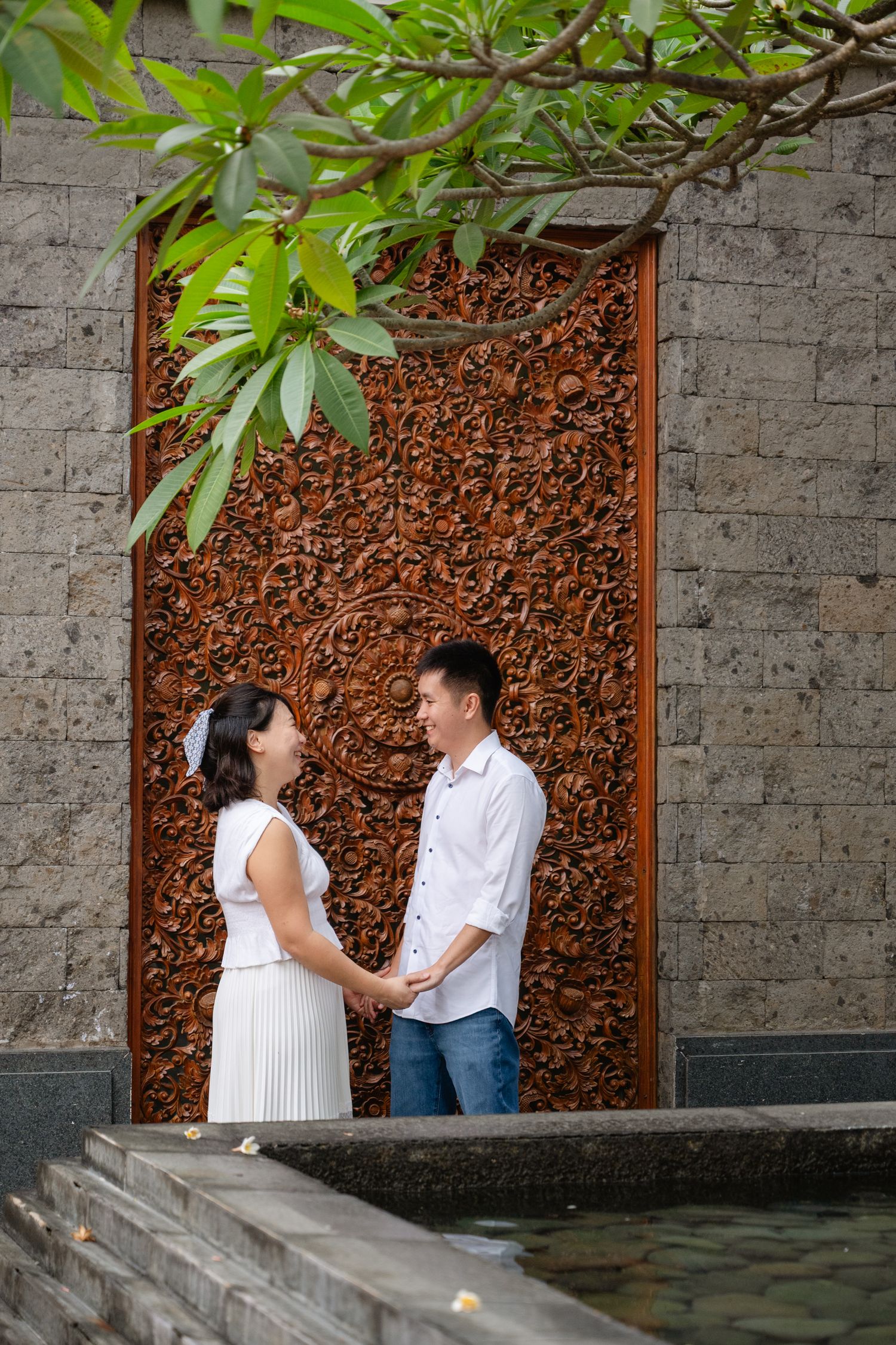 Couple holding hands in front of ornate Balinese carved wooden door with stone wall and tropical plants.