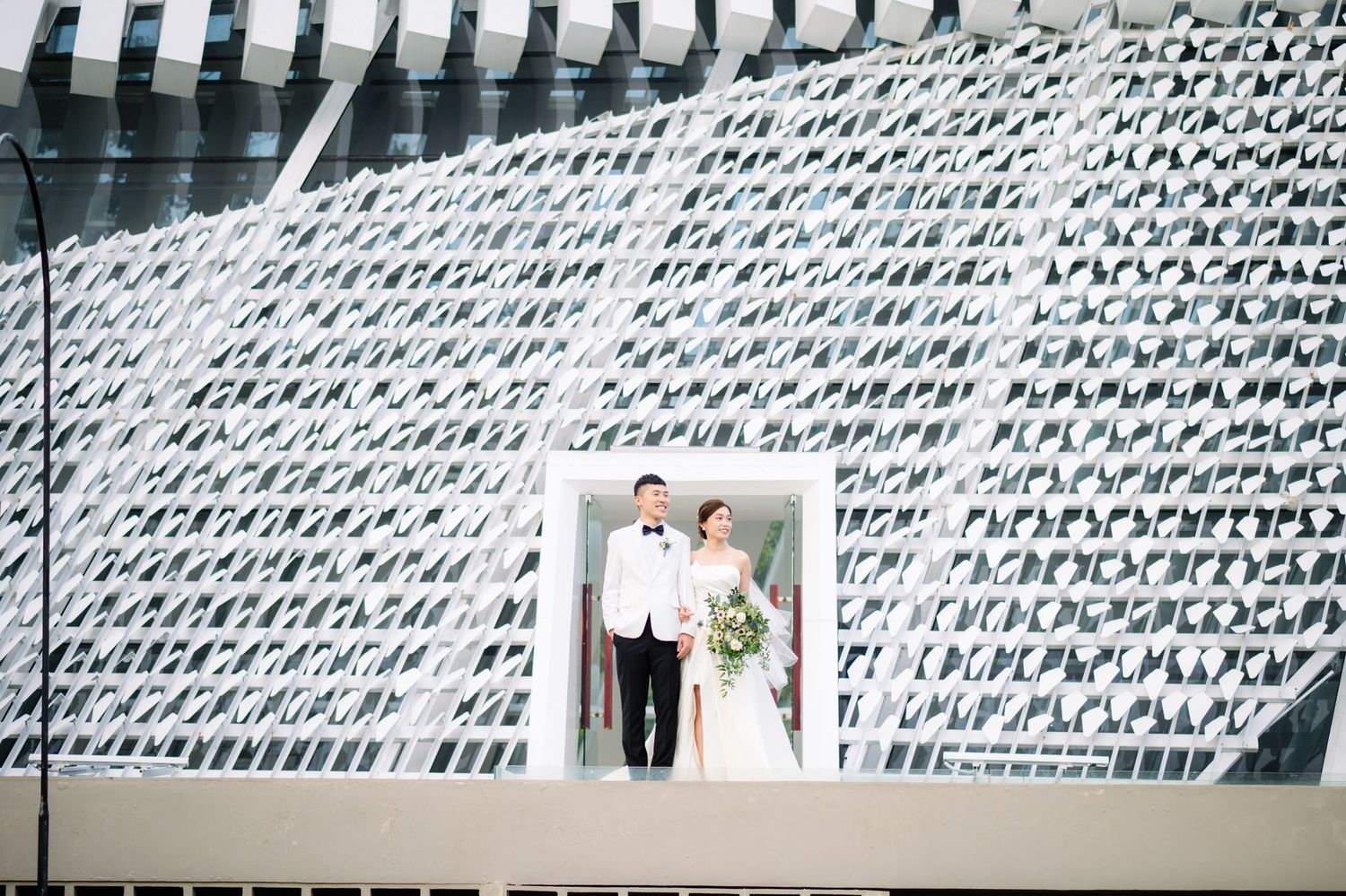 Bride in a white gown holding a bouquet and groom in a tuxedo pose in a doorway before a modern geometric patterned building facade.