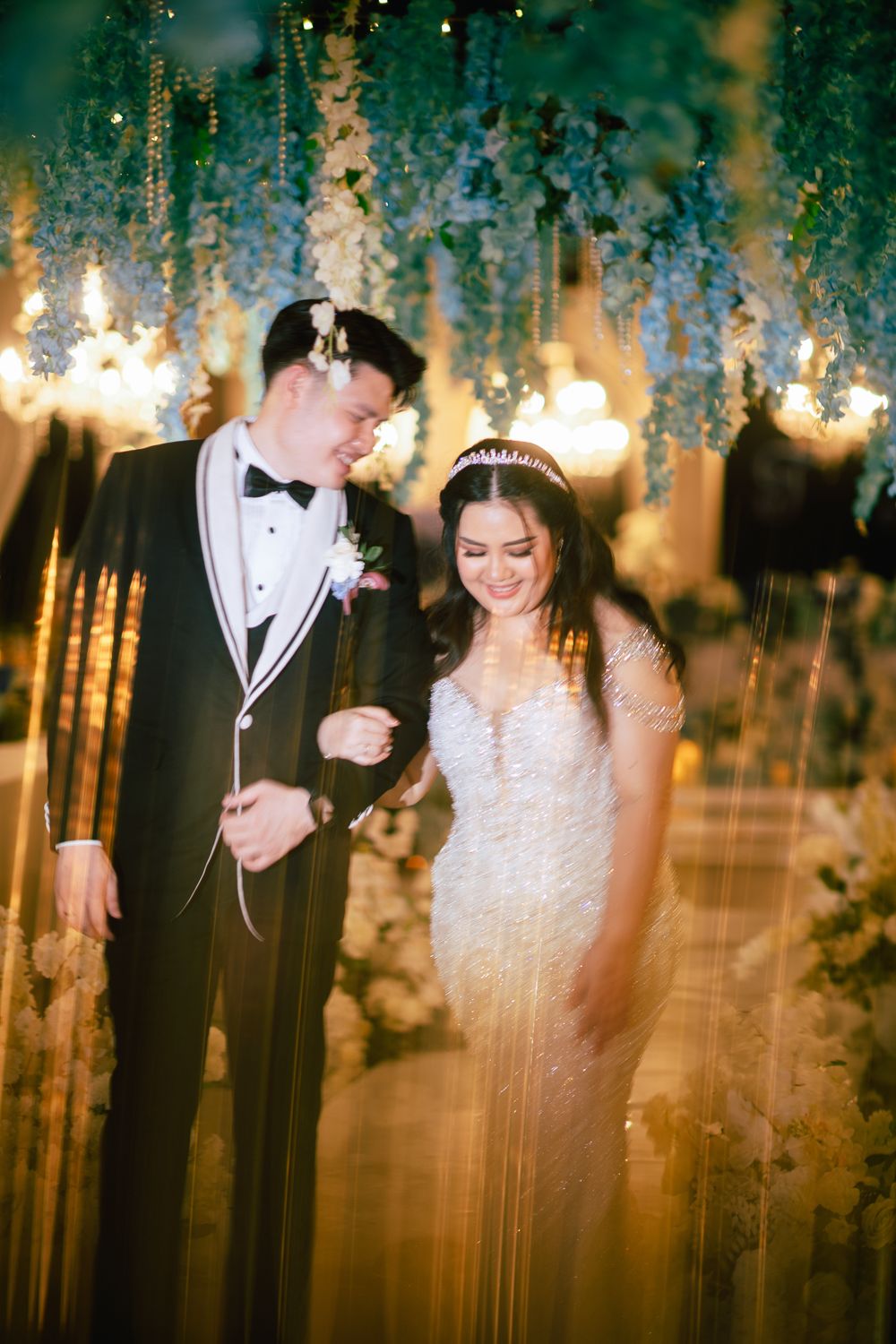 Bride in beaded gown and groom in black tuxedo share a joyful moment under floral chandeliers at elegant wedding reception.