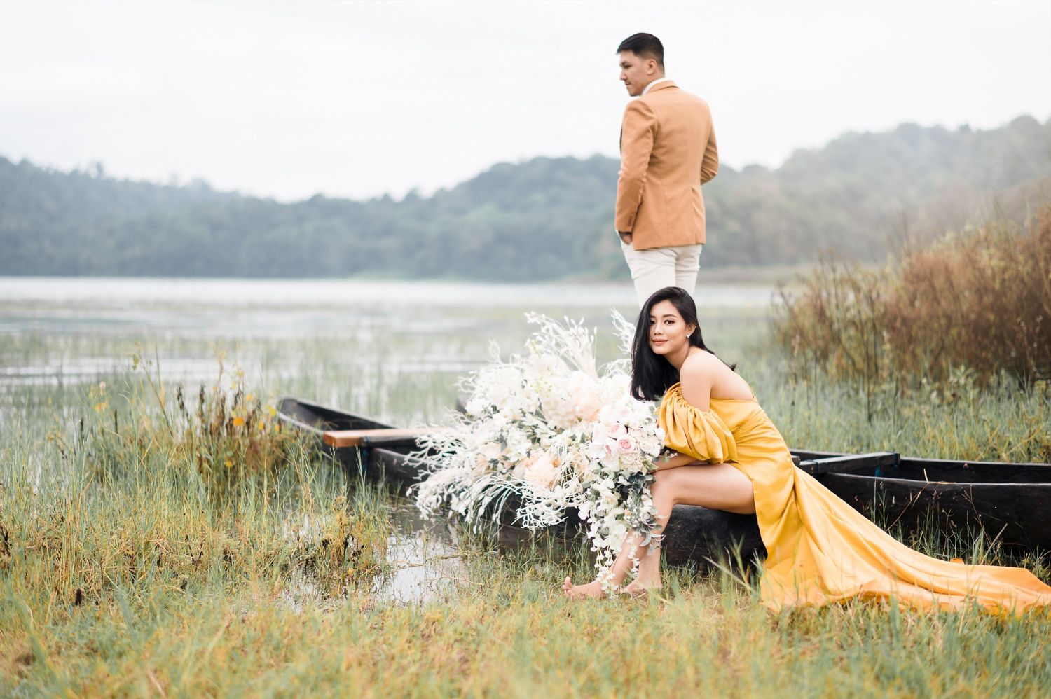 Couple in romantic outdoor photoshoot by a lake, woman in yellow gown with floral bouquet, man in tan blazer on a wooden boat.