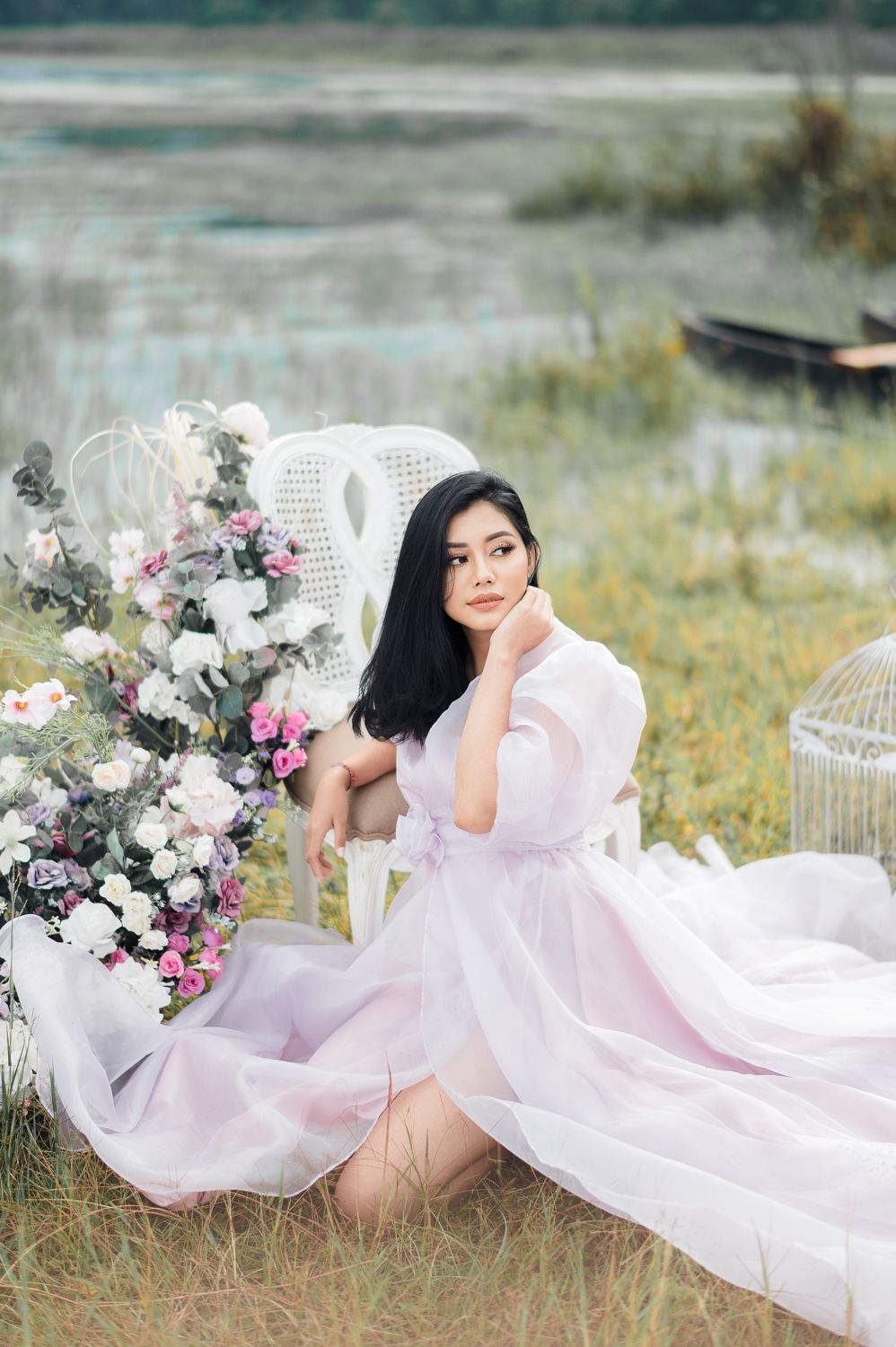 Young Asian woman in flowing pink dress sits gracefully outdoors near floral arrangement and white chair by a serene lake.
