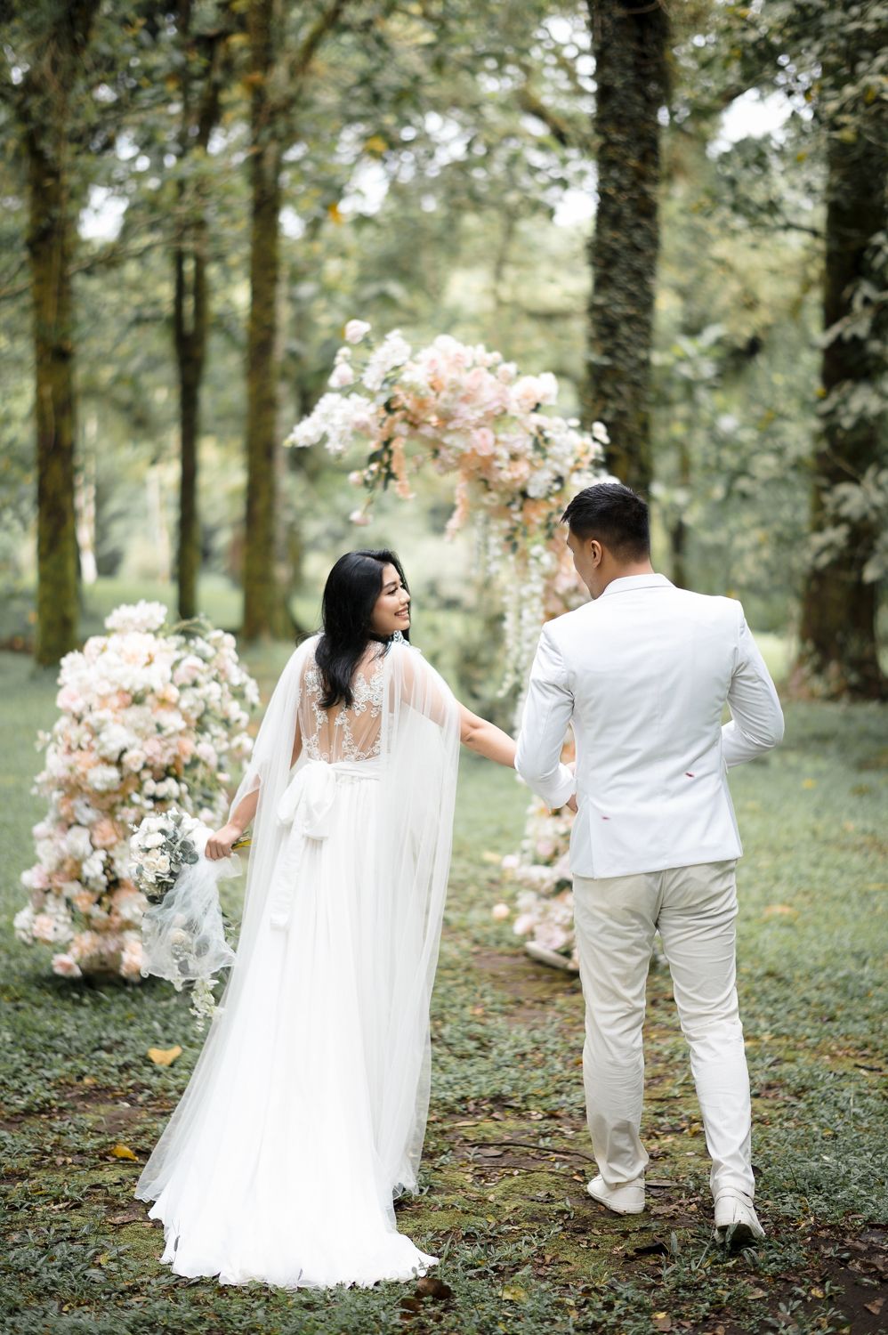 Bride in white cape gown holds groom's hand walking toward floral arch in lush forest wedding ceremony.