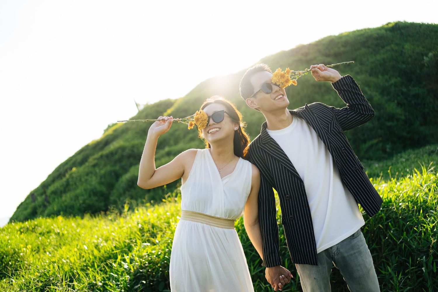 A happy couple holds wildflowers and laughs together on a sunny green hillside.