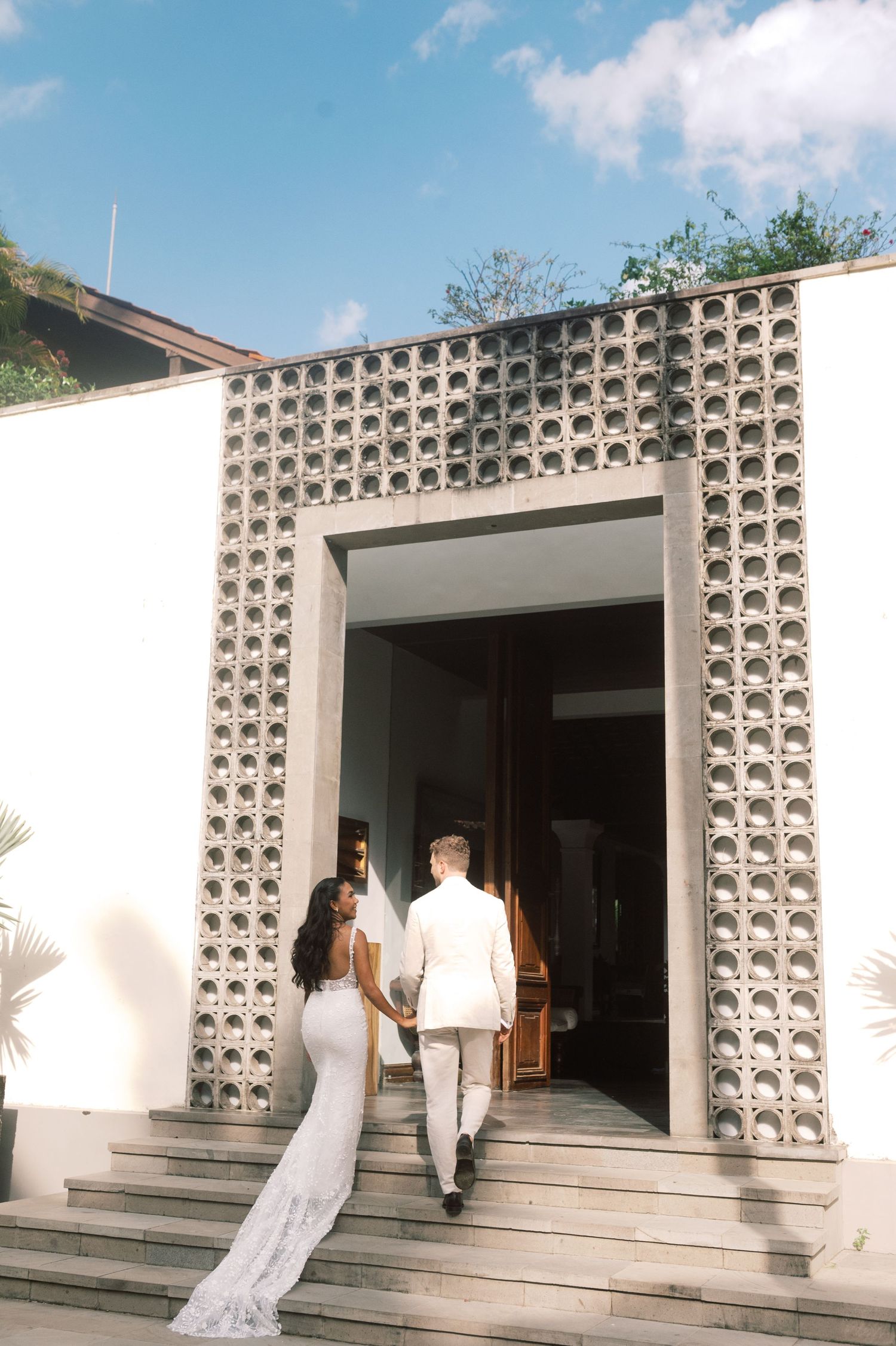 Bride and groom enter wedding venue through ornate concrete block archway under blue sky.