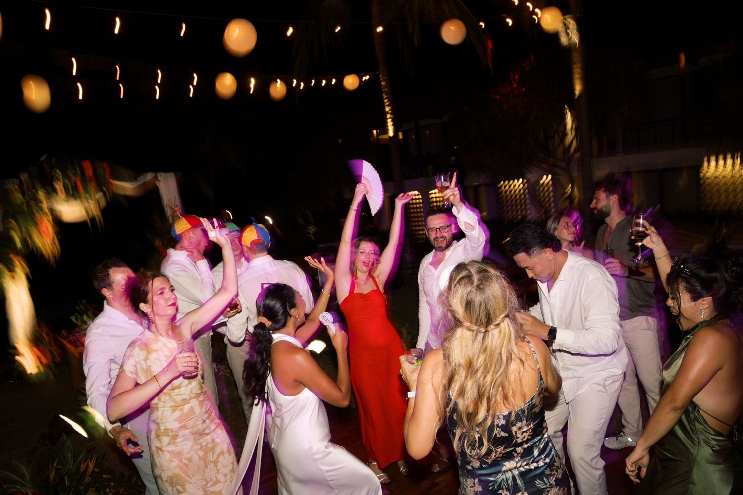 Bride, groom and guests dance joyfully together under string lights at a lively tropical wedding reception.