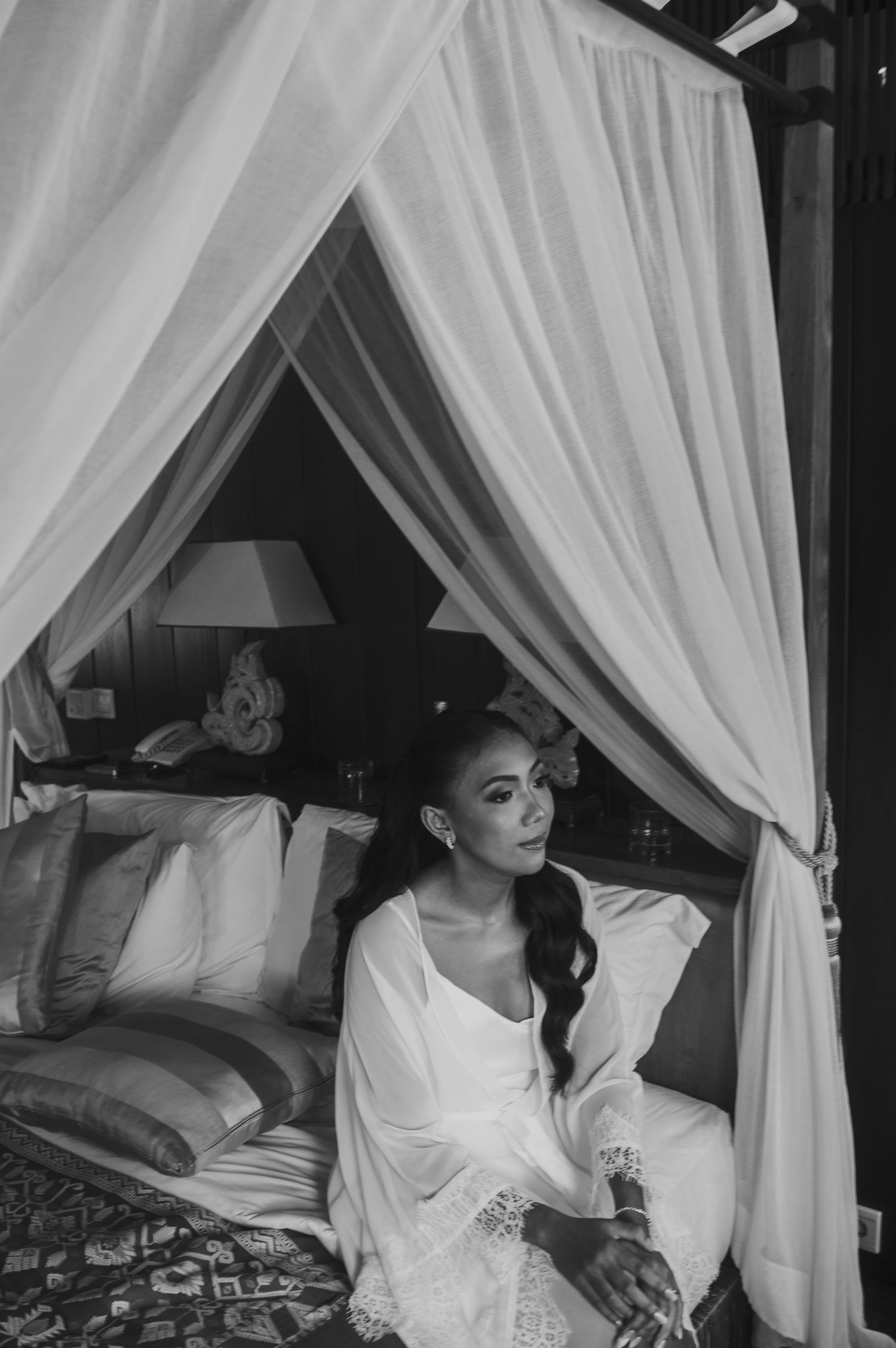 Bride in white robe sitting on a canopy bed in black and white wedding preparation photo.