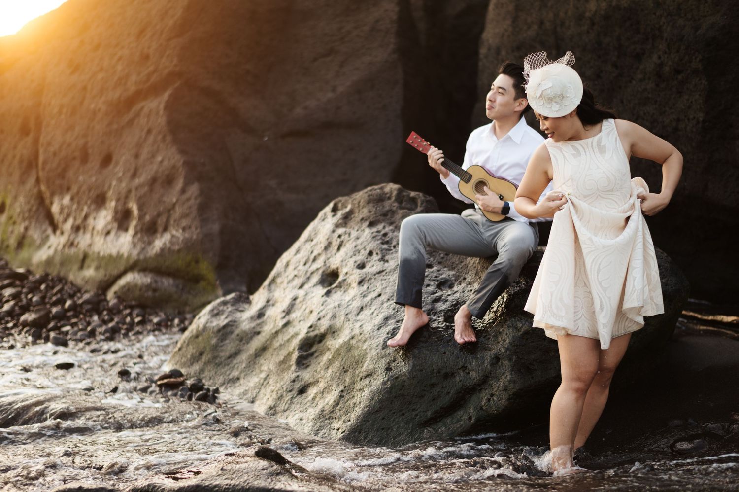 Couple on rocky beach at sunset, man playing ukulele while woman in white dress and hat stands nearby.