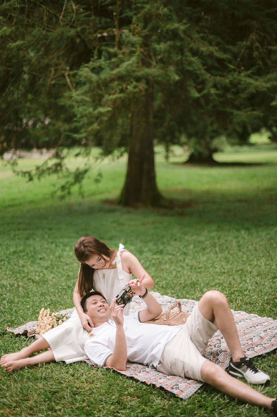 Couple enjoying a romantic picnic on a blanket in a lush green park, woman sitting while man lies down smiling.