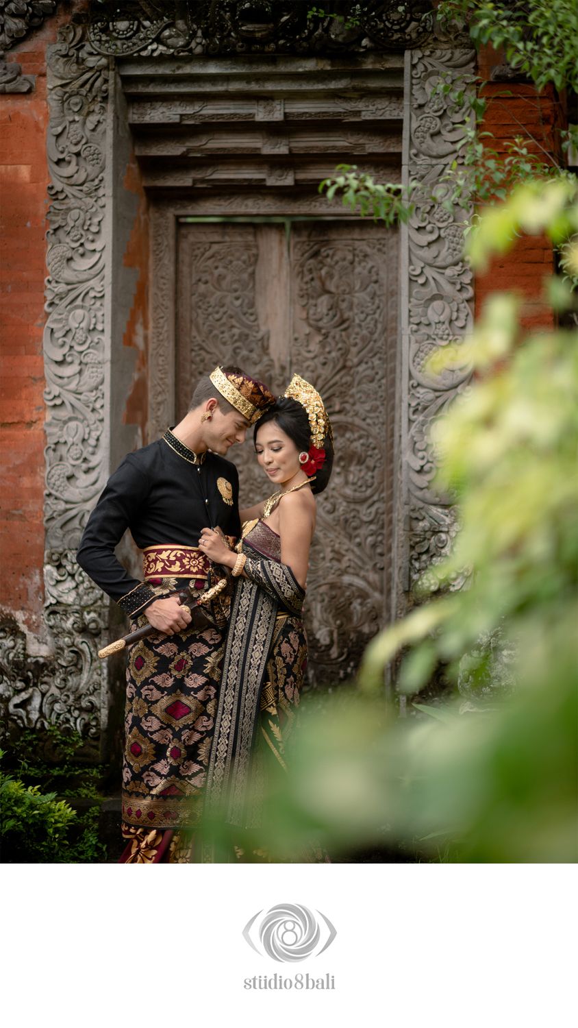 Balinese couple in traditional wedding attire posing before ornate stone temple door surrounded by lush greenery.