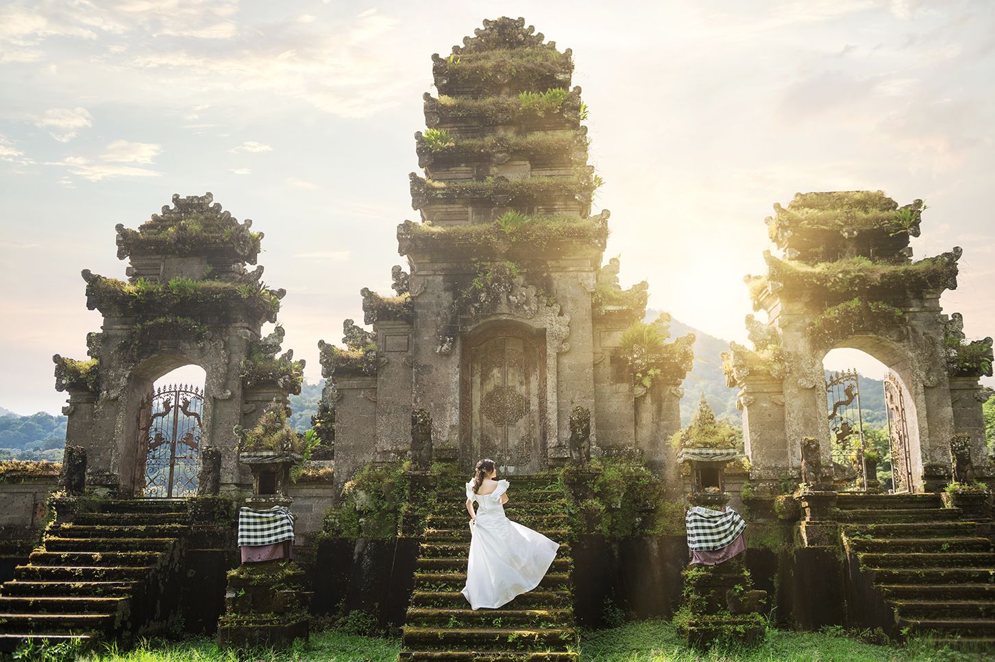 Couple in wedding attire posing at a moss-covered Balinese temple gate with sunlight streaming through at sunset.