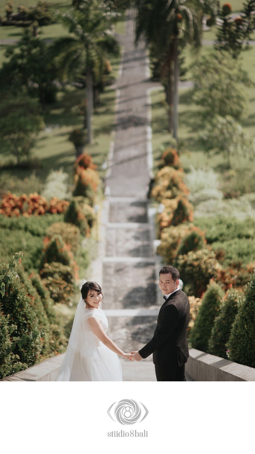 Bride and groom holding hands in a lush garden, posing for a romantic wedding photo at a scenic outdoor venue.