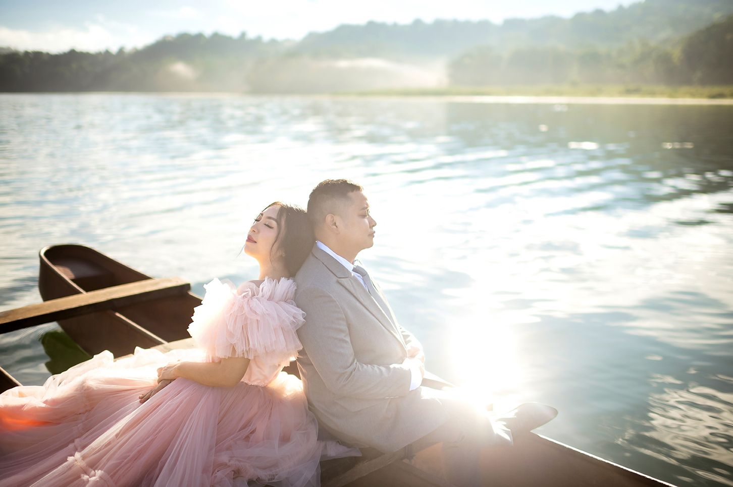 Couple in romantic pre-wedding shoot sitting back-to-back on a boat at a serene lake during golden hour.