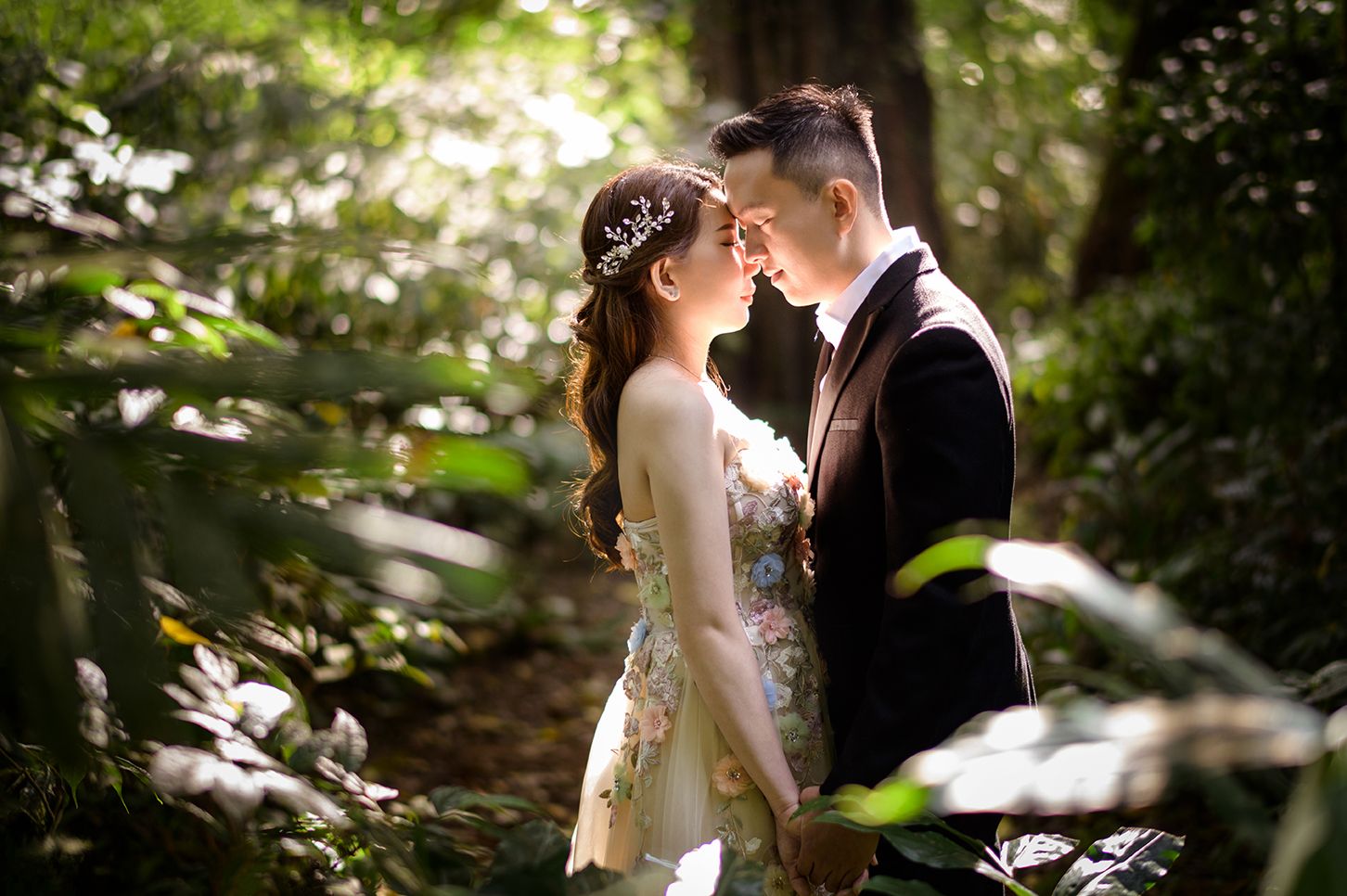 A couple in wedding attire shares a romantic moment in a sunlit forest, the bride wearing a floral gown and crown.