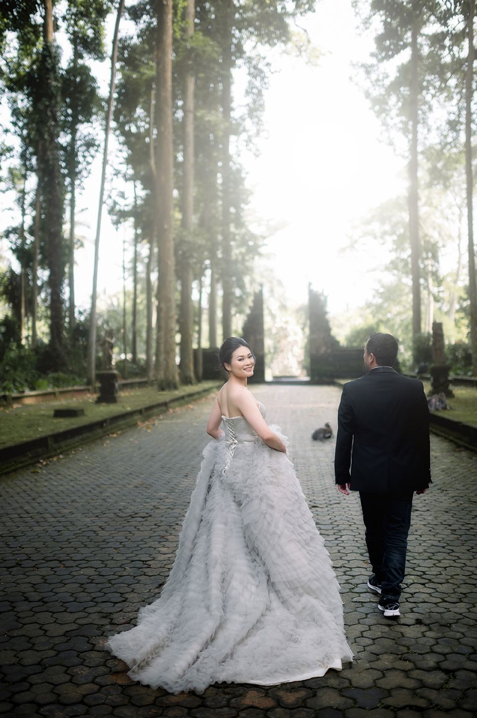 Bride in elegant white gown and groom in black suit walk cobblestone path through sunlit forest.