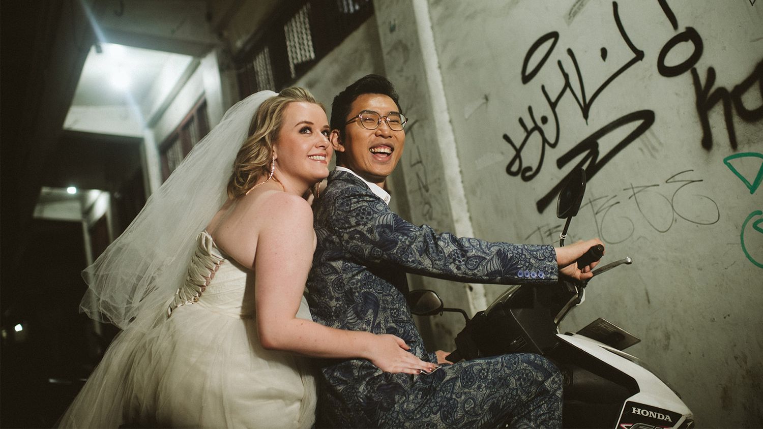 Bride in white gown and groom in blue floral suit riding a scooter together in an urban alley.