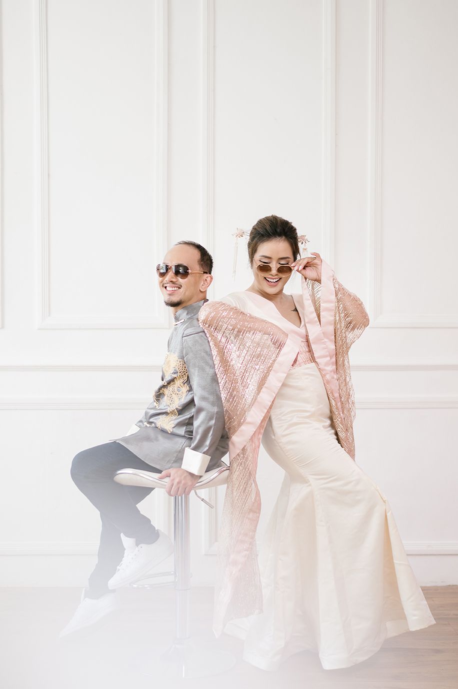 A stylish couple in traditional attire and sunglasses pose back-to-back on a chair in a bright white studio.