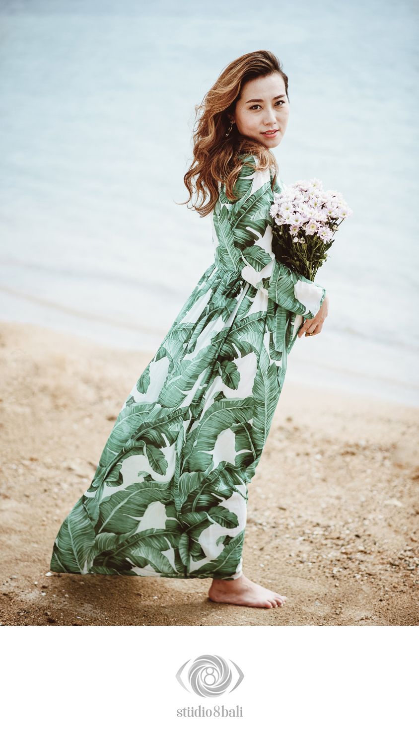 Woman in green tropical leaf maxi dress holds pink flowers on sandy beach, posing gracefully by the ocean.