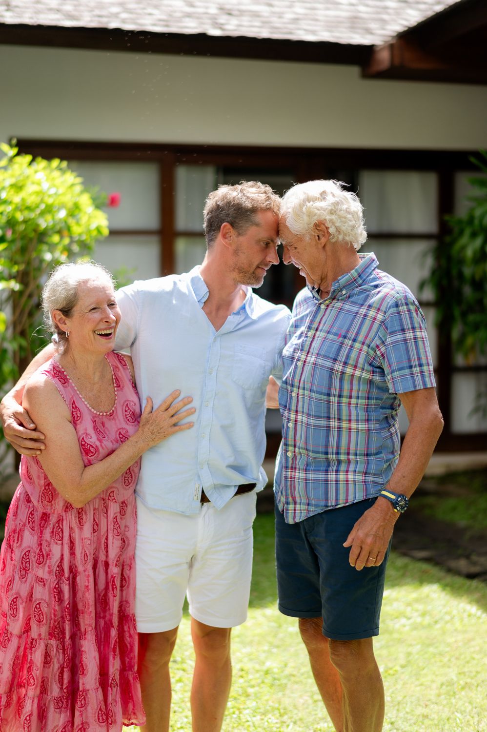 Senior couple and adult son sharing a warm embrace outdoors in sunny garden near a white house.