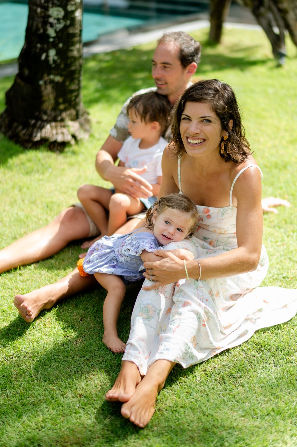 Family of four sitting on green grass near a pool, smiling mother holding toddler girl, father with young child behind.