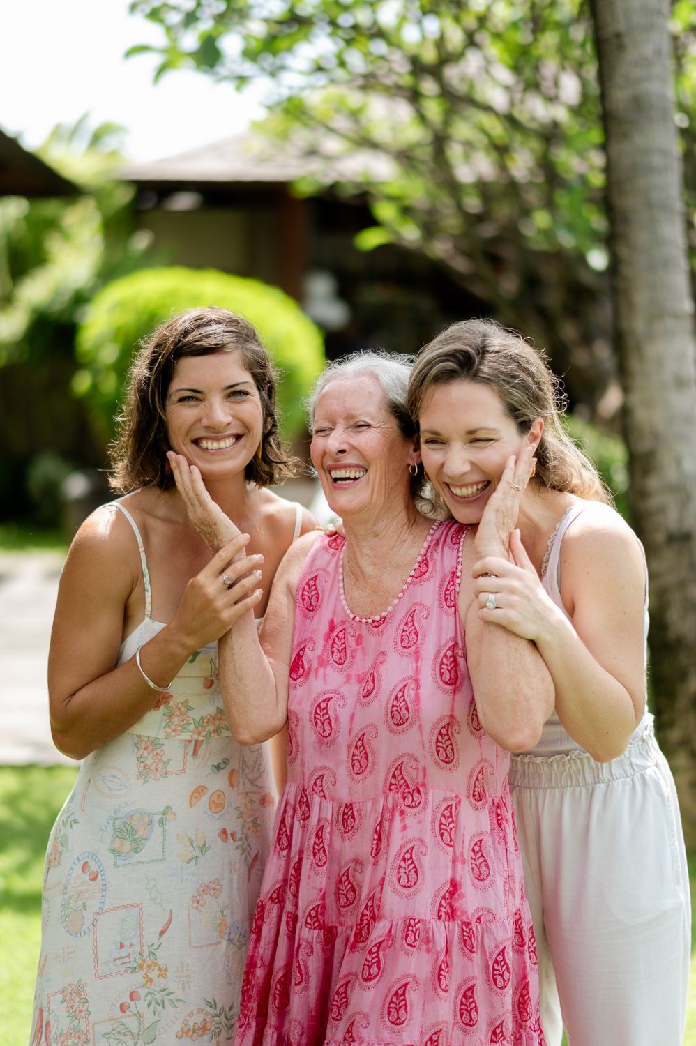 Three women smiling and laughing together outdoors, with lush greenery in the background.