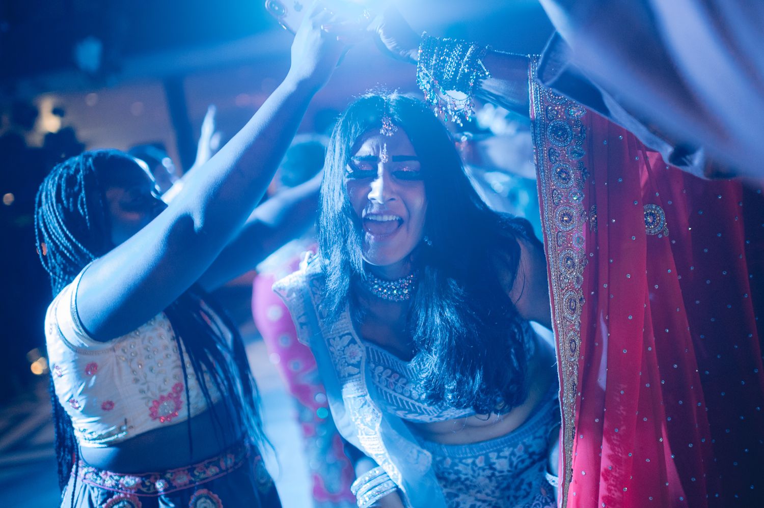 A bride in blue lehenga laughs joyfully while dancing with guests at a vibrant Indian wedding celebration.