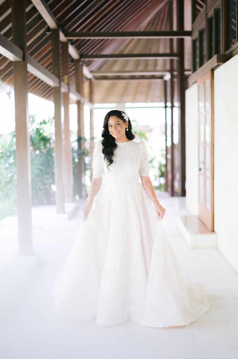 Bride in elegant white lace ball gown with elbow sleeves poses in a sunlit tropical venue corridor.