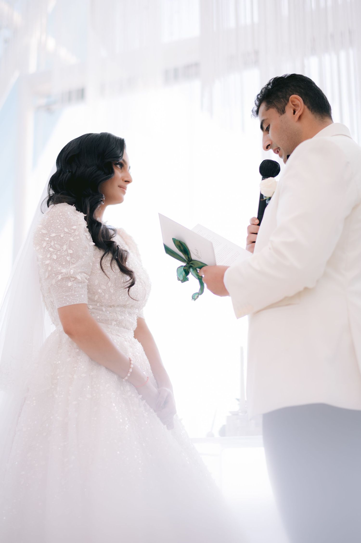 Bride and groom exchanging vows at a bright white wedding ceremony, groom reading from paper into microphone.