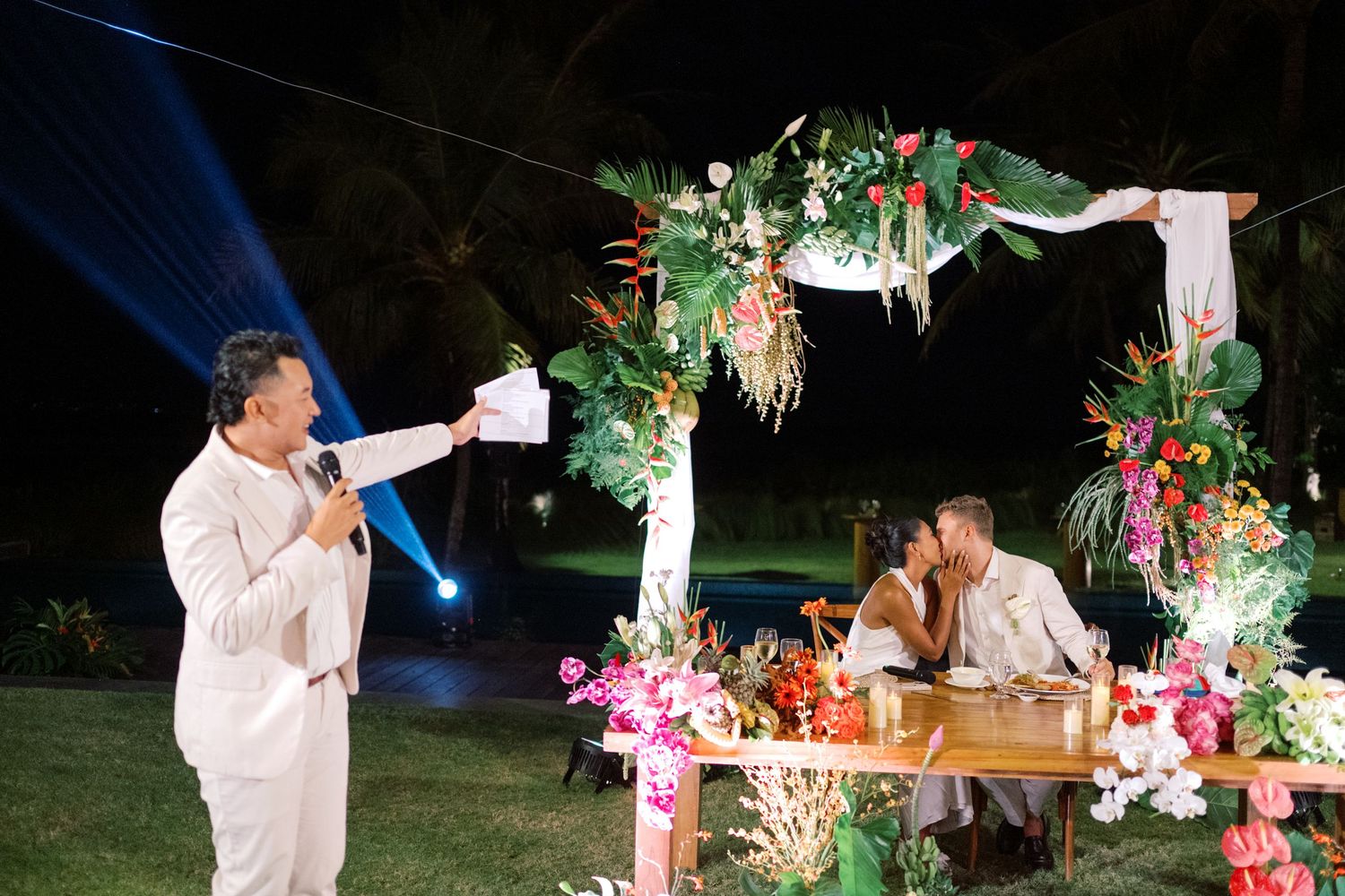 Wedding speaker at microphone while bride and groom kiss at floral decorated sweetheart table at night.
