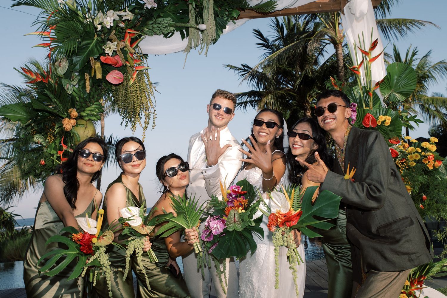 Bride and groom pose with bridesmaids in gold dresses under tropical floral wedding arch.