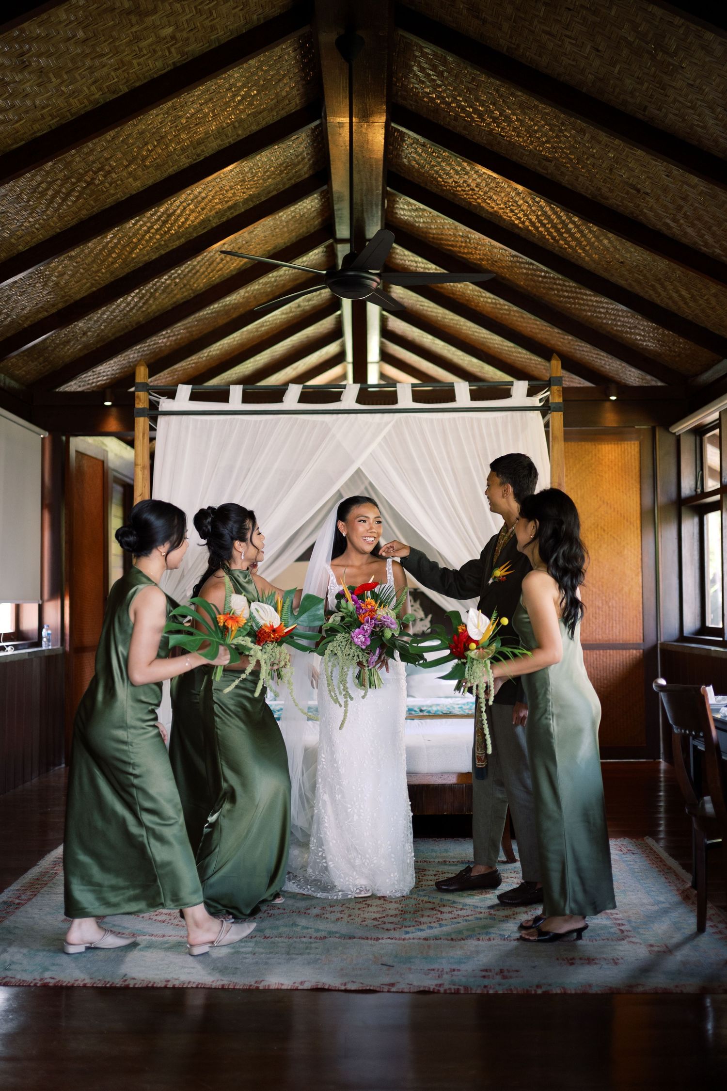 Bride in white gown with bridesmaids in olive green dresses holding tropical bouquets indoors.